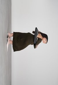 Moteris, vilkinti alyvuogių žalia suknelė ir baltus kulnus, laiko didelį juodą rankinį krepšį, stovėdama priešais paprastą šviesiai pilką fono.