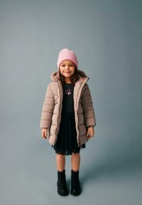 Beżowy płaszcz ocieplany z kapturem, w zestawieniu z czarną sukienką z grafiką serca, różowym dzianinowym beanie i czarnymi botkami za kostkę.
