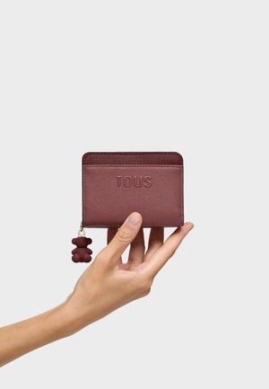 Mano sosteniendo una pequeña cartera de cuero marrón vino con el logo "TOUS" en relieve y un tirador de cremallera en forma de oso contra un fondo liso.