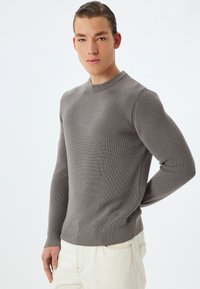 Grijze gebreide trui met een ronde hals, gestructureerde stof en geribbelde zoom. Model draagt crèmekleurige broek, handen achter de rug.