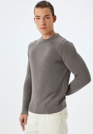 Suéter gris de punto con cuello redondo, tejido texturizado y dobladillo acanalado. Modelo viste pantalones color crema, con las manos detrás de la espalda.