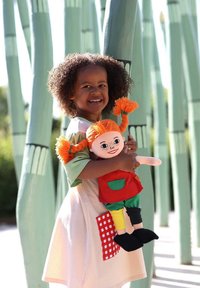 Child with curly hair holds a fabric doll dressed in a red top, green pants, and black shoes. The dress features a beige striped design with a checkered pocket.