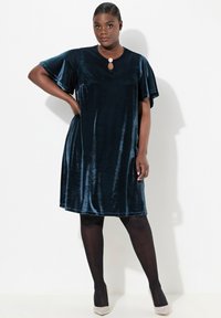 Tumma sininen samettimekko, jossa on lyhyet liehuvat hihat ja avok oharuksella kaula-aukossa. Sileä pinta, polveen ulottuva, yhdistetty mustiin sukkahousuihin ja korkokenkiin.