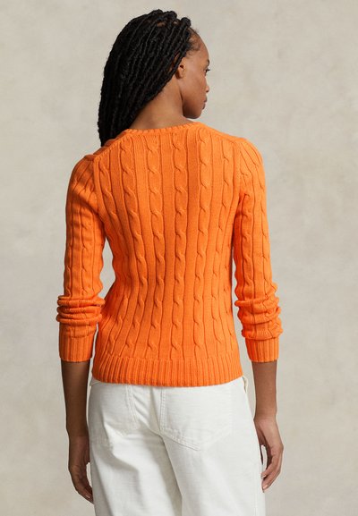 Pull en tricot orange avec un motif en câble, coupe ajustée, manches longues et bords-côtes aux poignets et à l'ourlet, porté avec un pantalon blanc.