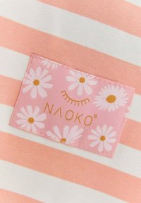 Pruhovaná látka v ružovej a bielej farbe s ružovou nášivkou s bielymi sedmikráskami a textom "NAOKO" v zlatej farbe. Hladká, ľahká textúra.