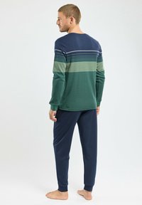 Homme debout pieds nus portant un haut à manches longues rayé vert et marine et un pantalon marine uni, tourné le dos contre un fond uni.