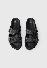 Black leather sandals with double buckle straps, white stitching, and round studs, labeled "TOGA VIRILIS" on the footbed.