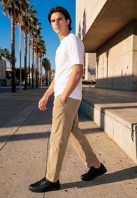 Jeune homme marchant sur un trottoir urbain bordé de palmiers, portant un t-shirt blanc, un pantalon beige et des baskets noires en pleine lumière du jour.