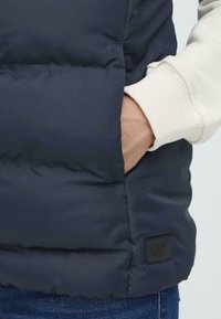 Gros plan sur une main dans une poche d'une doudoune noire, avec une manche de couleur claire visible. Une étiquette de logo est visible sur le bord inférieur de la veste.