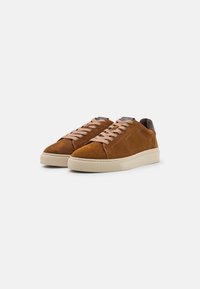 Braune Wildledersneaker haben eine cremefarbene Gummisohle, beigefarbene Schnürsenkel und einen dunkelbraunen Lederakzent an der Ferse. Minimalistisches Design.