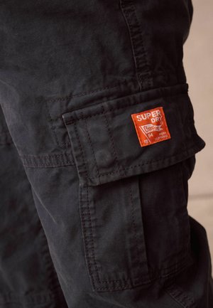 Pantaloni cargo neri realizzati in un tessuto testurizzato, dotati di un'etichetta arancione "Superdry" sulla tasca laterale e dettagli di cucitura in vista.