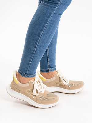 Person wearing light brown and beige sneakers with white soles and laces, paired with blue skinny jeans, standing on a white surface.