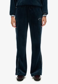 Navyblaue Velour-Hosen mit weitem Bein, seitlichen Streifen und dem eingestickten "Superdry"-Logo auf dem linken Oberschenkel. Weiche Textur und elastischer Bund.