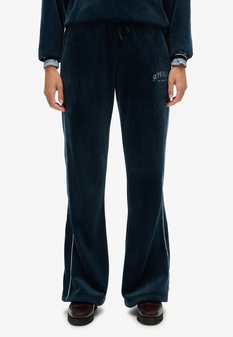 Navyblaue Velour-Hosen mit weitem Bein, seitlichen Streifen und dem eingestickten "Superdry"-Logo auf dem linken Oberschenkel. Weiche Textur und elastischer Bund.