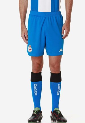 Athlète masculin portant un short de football bleu Deportivo avec écusson, des chaussettes bleues avec le texte "DEPOR", des bandes de chaussettes noires et des crampons noirs.