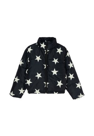 Jeune personne portant une doudoune noire avec des étoiles blanches, un jean gris clair à jambes larges et des bottes noires, debout avec une main dans la poche.