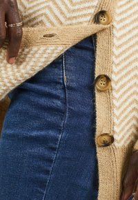 Cardigan a zigzag tan e bianco con tre bottoni marroni, indossato sopra jeans in denim blu scuro. Texture morbida.