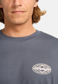 Graues Baumwoll-T-Shirt mit Rundhalsausschnitt, das ein weißes ovales Logo mit Text zeigt. Glatte Textur, lässige Passform und kurze Ärmel.