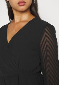 Femme portant une robe portefeuille noire avec des manches longues en voile présentant un motif chevron texturé, des boucles d'oreilles créoles et un maquillage naturel.