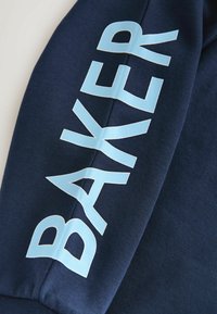 Temno modra majica z gladkim tekstilom; na levem rokavu je velik svetlo moder napis "BAKER".