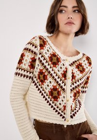 Frau in einer cremfarbenen Häkeljacke mit geometrischen Mustern in Orange, Braun und Oliv, mit geknöpftem Vorderteil, kombiniert mit braunen Hosen.