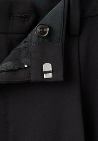 Tessuto nero con una texture liscia presenta un bottone e una fibbia metallica. È visibile una cerniera, che mette in risalto gli elementi di design funzionali del capo.