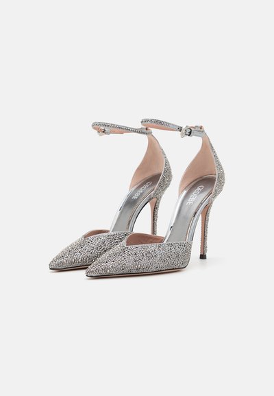 Zapatos de tacón alto plateados con punta afilada, cubiertos de brillantes strass. Cuentan con correas en el tobillo y una plantilla metálica.