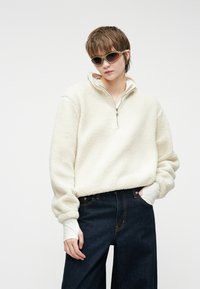 Pullover in pile color crema con colletto alto e zip a metà, abbinato a jeans a gamba larga blu scuro e maniche bianche sovrapposte.