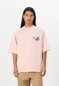 Lys rosa oversized t-skjorte med korte ermer, med et sirkulært grafisk design av en fisk på venstre brystområde. Mykt bomullsstoff.