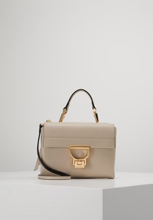 Sac à main en cuir beige avec un fermoir doré, une poignée supérieure et une bandoulière noire amovible sur un socle blanc.