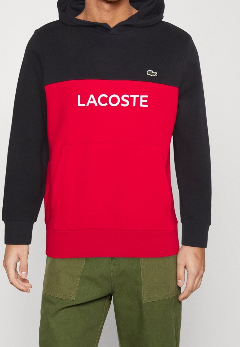 Hoodie med röd kropp och svarta ärmar, med vit "LACOSTE"-logotyp och kenguruficka, tillverkad av texturerad tyg.