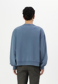 Sudadera azul con un corte relajado, cuello redondo y hombros caídos. Textura suave con puños y dobladillo acanalados. Llevada sobre pantalones grises texturizados.