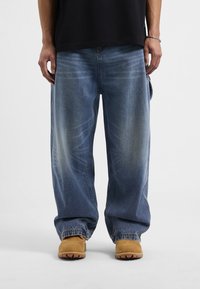 Luźne niebieskie dżinsy charakteryzujące się wyblakłymi akcentami i swobodnym krojem, w połączeniu z jasnobrązowym obuwiem i czarną koszulą.