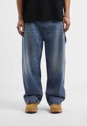 Baggy blue denim jeans feature faded accents and a relaxed fit, paired with tan footwear and a black shirt.