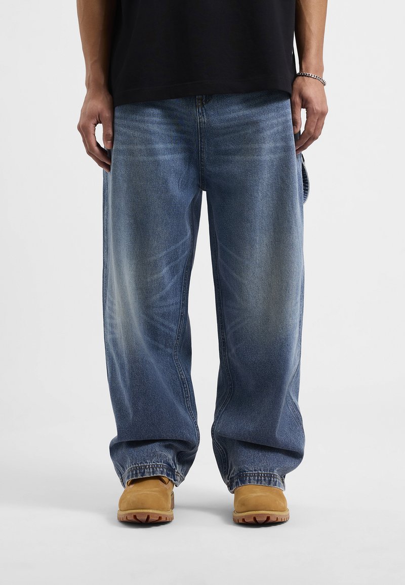 Luźne niebieskie dżinsy charakteryzujące się wyblakłymi akcentami i swobodnym krojem, w połączeniu z jasnobrązowym obuwiem i czarną koszulą.