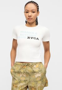 Jeune femme aux cheveux tressés portant un t-shirt blanc "THANKS 4 NOTHING CLUB RVCA" et un short vert à imprimé tropical, les mains dans les poches.