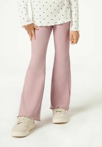 Pantalones acampanados de canalé rosa claro con una superficie texturizada. Los puños están ligeramente deshilachados. Combinados con zapatillas deportivas beige.