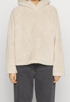 Personne portant un pull à capuche beige et moelleux associé à un jean ample taille haute gris foncé, debout devant un fond uni.