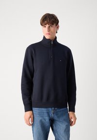 Navy blue pullover sweatshirt with a high collar and half zip closure. Features a subtle logo on the chest and ribbed hem and cuffs.