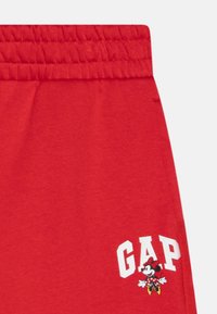 Rde bombažne trenirke z elastičnim pasom, ki imajo bel "GAP" logo in grafiko Mikija Miške v spodnjem levem kotu.