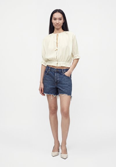 Femme debout portant un chemisier crème noué devant à manches bouffantes, un short en jean coupé avec des ourlets effilochés et des talons blancs pointus.