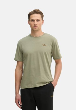 Uomo che indossa una maglietta verde chiaro a maniche corte e pantaloni neri con un logo alato dorato sul petto e sulla coscia, in piedi con una mano in tasca.