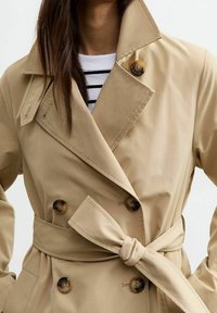 Mujer vestida con un trench coat beige de doble botón con botones de carey y cinturón atado en la cintura, sobre una blusa de rayas blanca y negra.