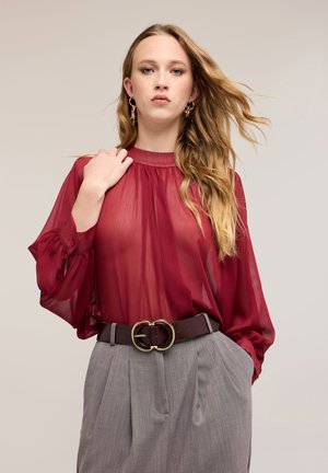 Jonge vrouw met lang blond haar draagt een doorschijnend rood blouse, grijze broek met hoge taille en een donkere riem met een gouden dubbele ringgesp.