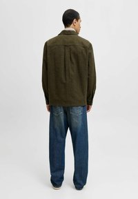 Mand set fra ryggen iført olivenfarvet jakke og løse blå jeans mod en ensfarvet lys baggrund.