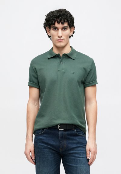 Polo en coton vert avec col, manches retroussées et petit logo sur la poitrine, assorti à un jean bleu foncé et une ceinture noire.