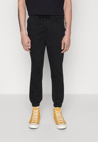 Schwarze Jogginghosen mit elastischen Bündchen, Kordelzugbund und zwei vorderen Aufsatztaschen. Tragen mit leuchtend gelben High-Top-Sneakern.