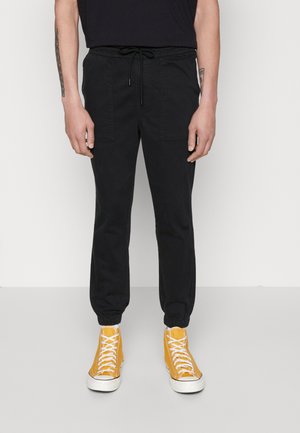 Pantaloni jogger neri con polsini elasticizzati, cintura con cordino e due tasche anteriori a toppa. Indossati con sneakers alte gialle brillanti.