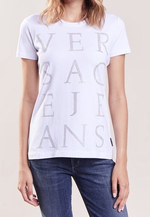 Femme portant un T-shirt blanc à manches courtes avec des lettres perforées formant le mot "VERSACE JEANS" et un jean en denim bleu.