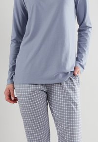 Top a maniche lunghe di un blu chiaro abbinato a pantaloni a quadri nei colori blu, bianco e lavanda. Tessuto morbido, vestibilità comoda e tasche laterali visibili.